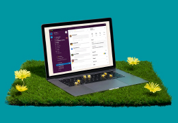 Open laptop displaying a Slack workspace interface sitting on a patch of green grass with yellow dandelions against a teal background.