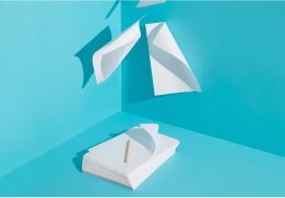 A stack of paper with a pencil on top and sheets floating against a bright blue backdrop.